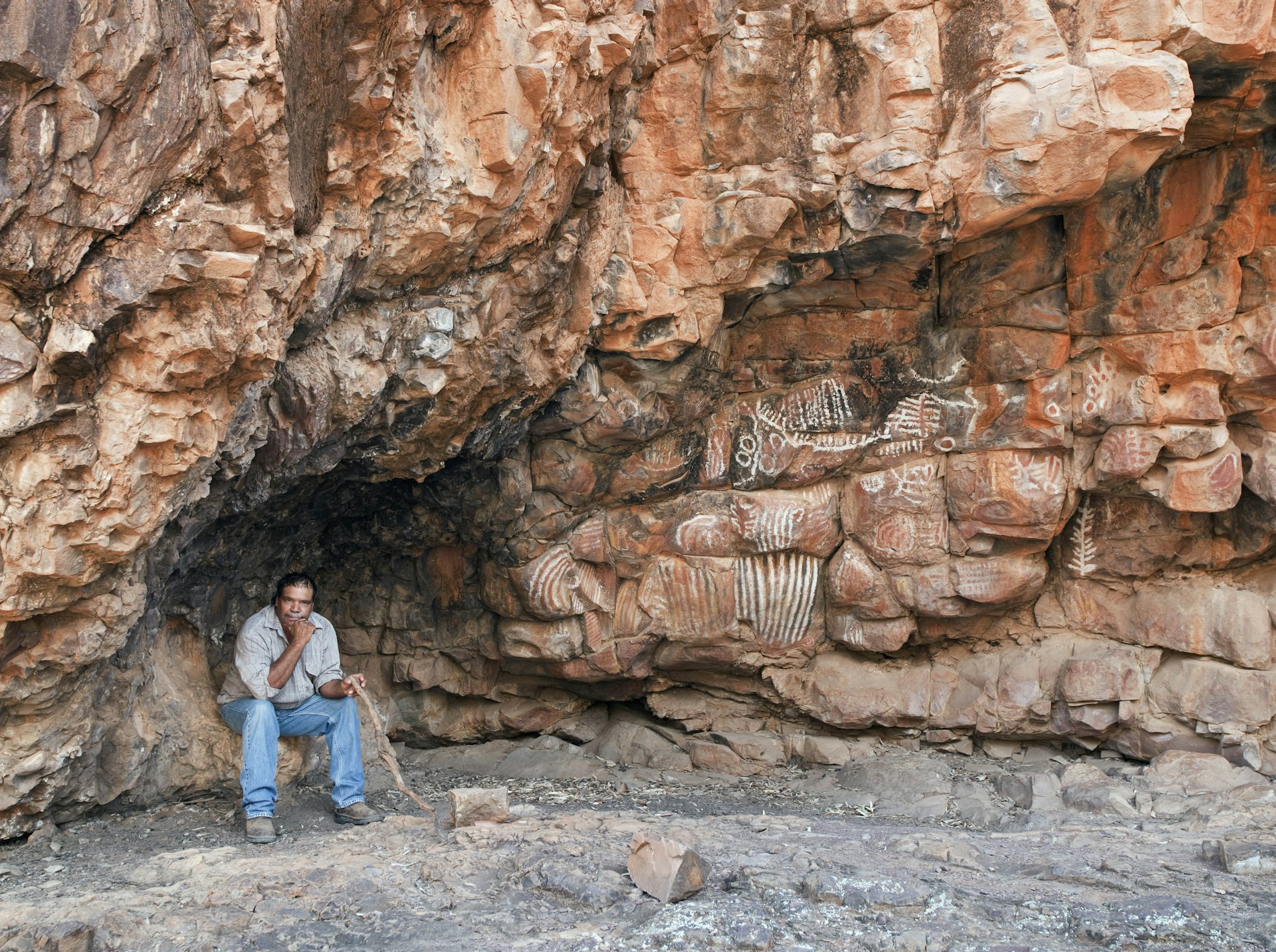 An Aboriginal man sits at the base of a large rock face daubed with rock art drawings