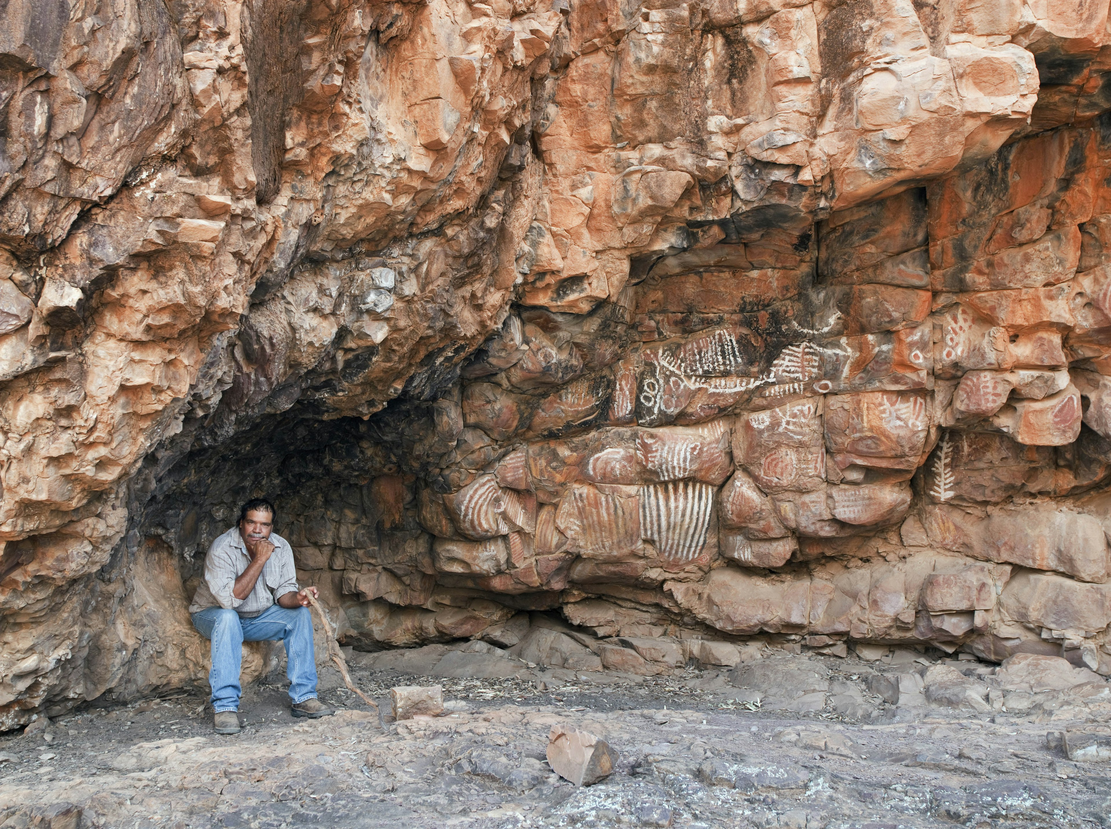 An Aboriginal man sits at the base of a large rock face daubed with rock art drawings