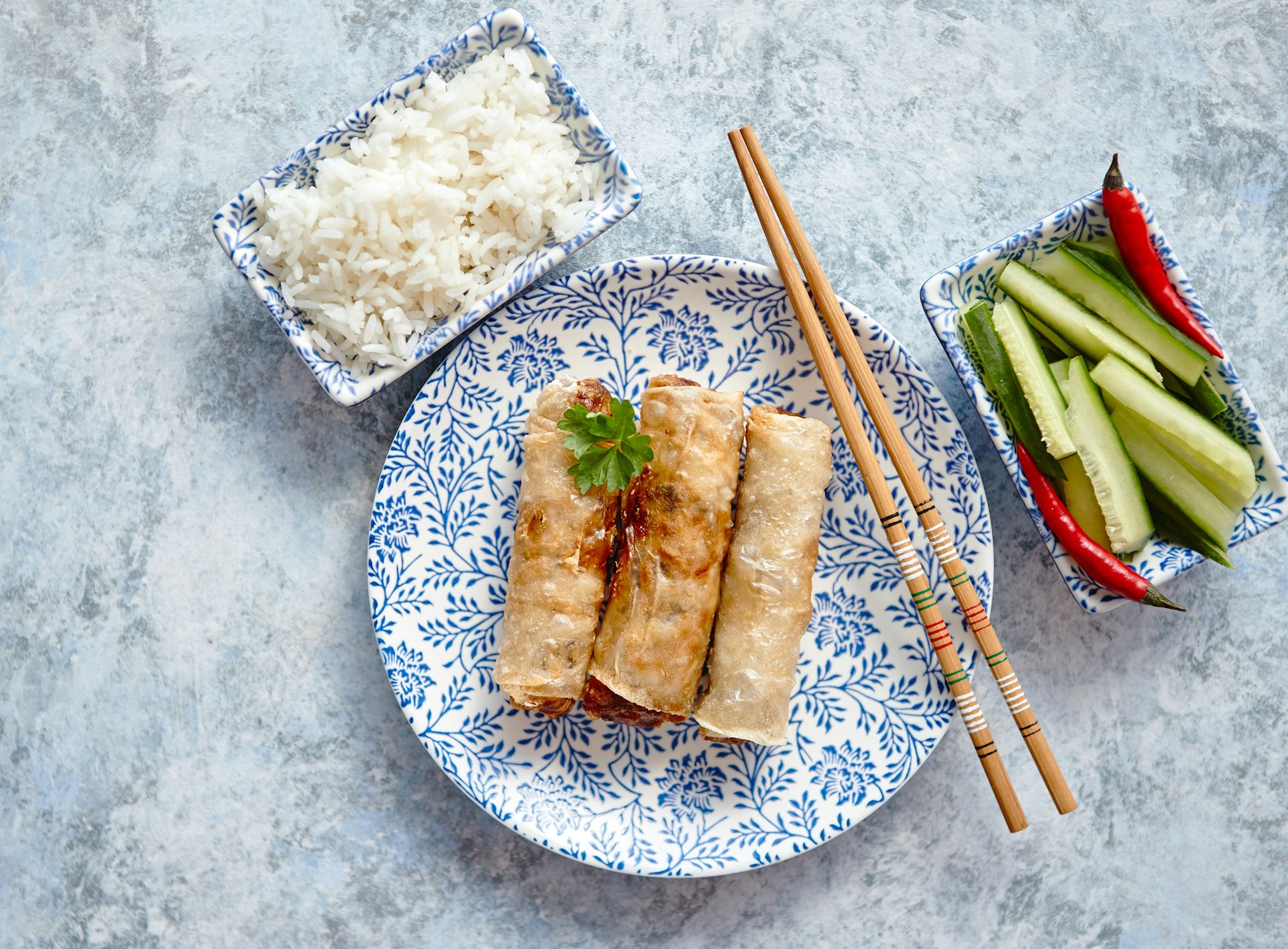 A top-down view of a handful of deep fried spring rolls placed on beautiful plate with chopsticks, and boiled rice on the side.