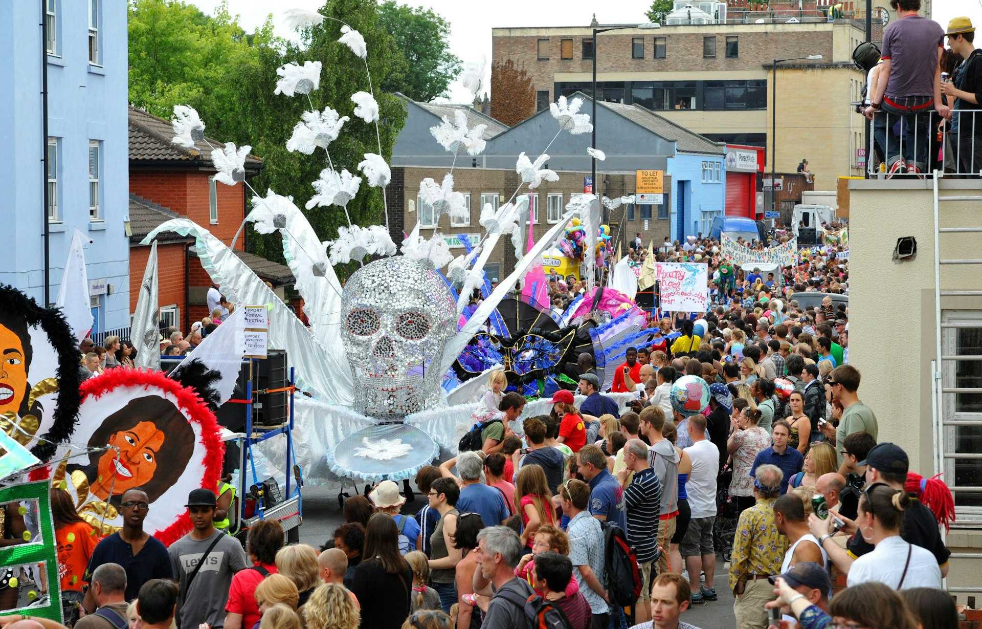 A giant float shaped like a huge skull with feathers goes down a street. People line the street watching the carnival procession