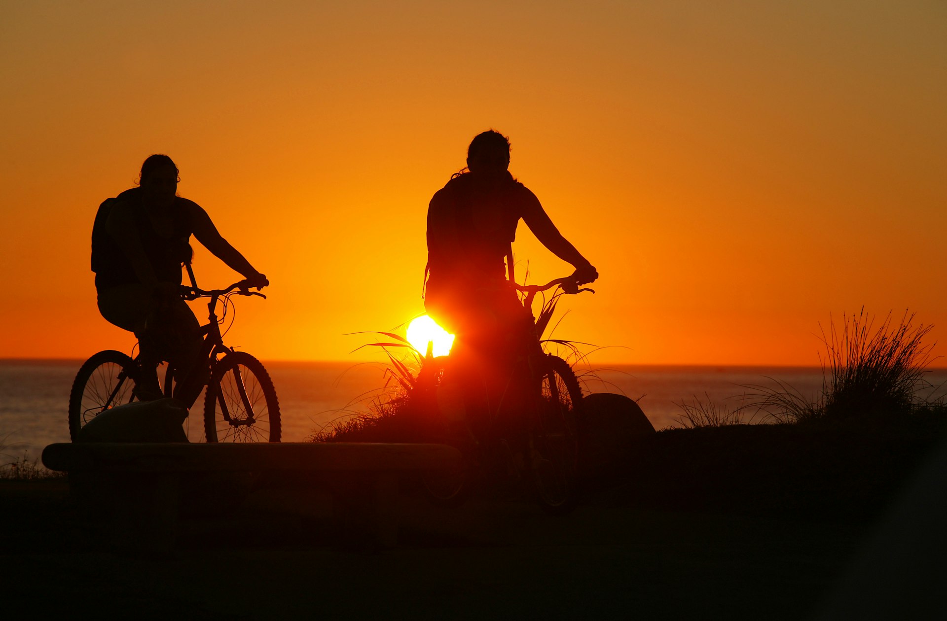 Two people in silhouette ride on bikes during sunset near a large body of water. 