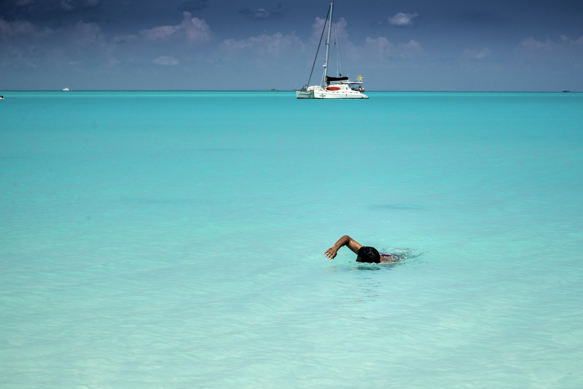 Man swimming to shore in perfectly clear turquoise-colored water. A catamaran sailboat at anchor is in the background.