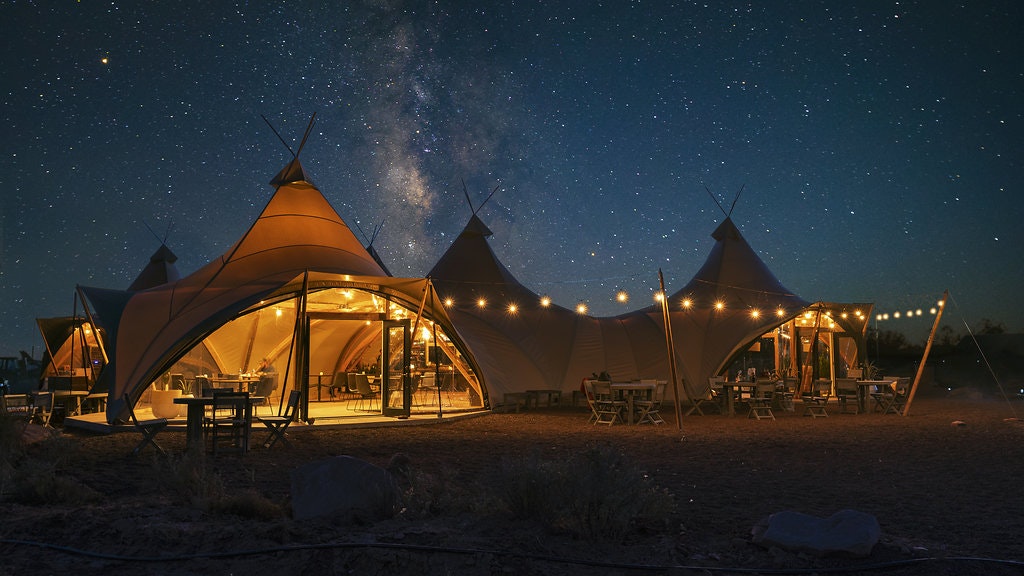 A safari-style tent with a large white canopy and string lights hanging outside. It's night and the stars are brightly shining down, filling the sky