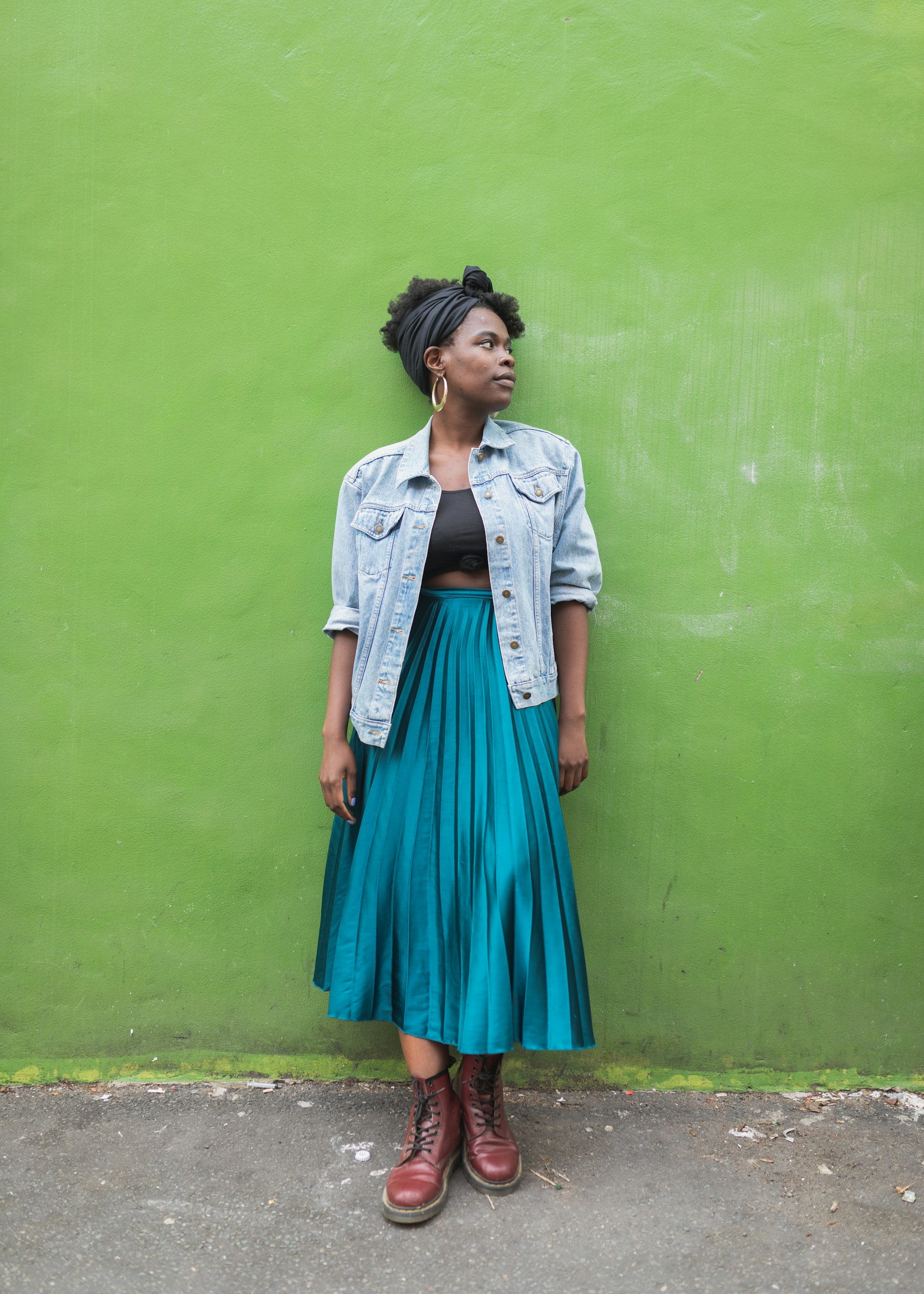 A black woman wearing a long skirt and denim jacket stands against a green wall
