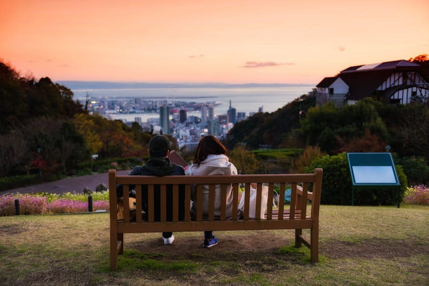 Sweet young couple sitting on wooden chair seat to enjoy aerial view of Kobe Nunobiki Herb Gardens and skyline cityscape with twilight sky at dusk in Japan