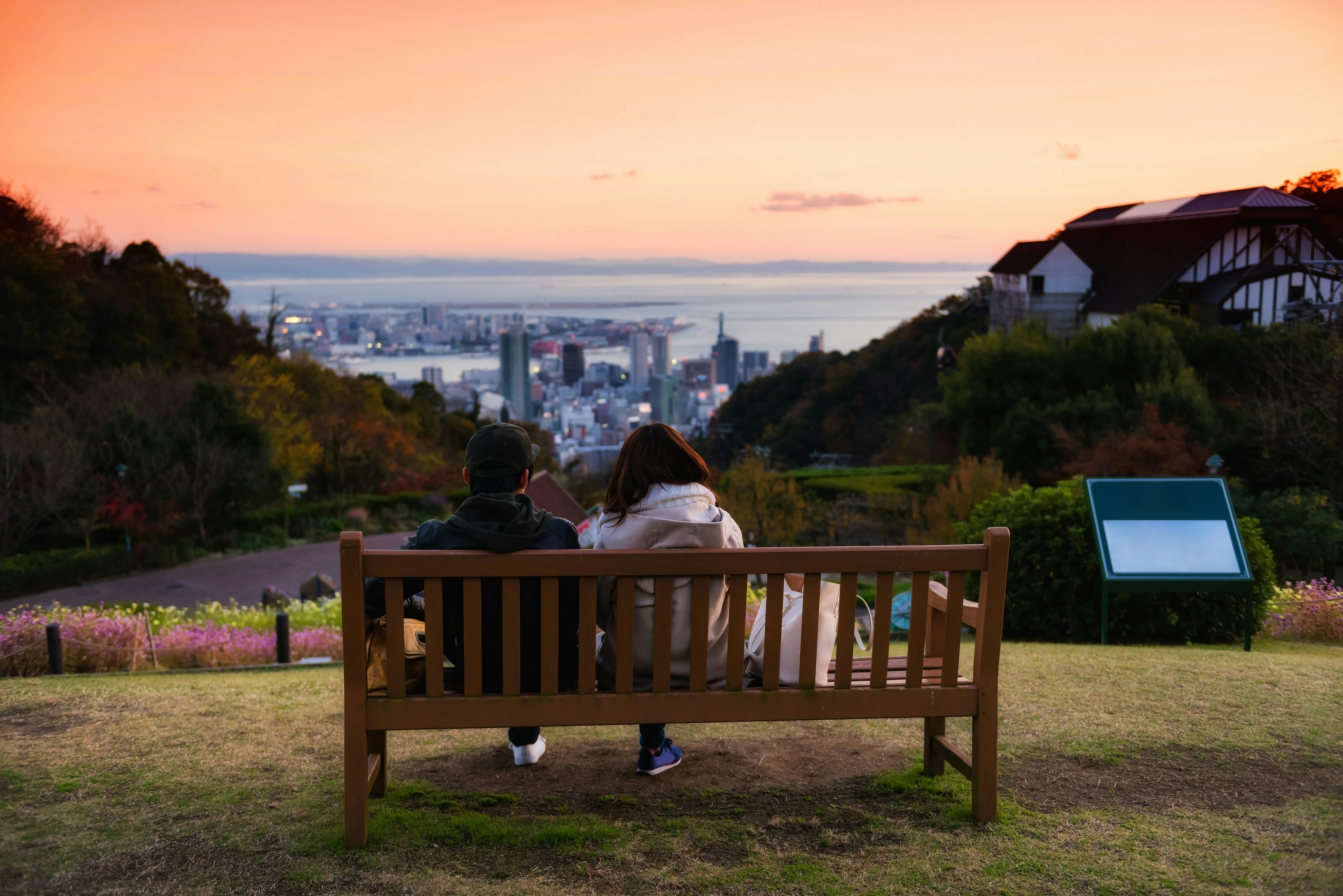 Sweet young couple sitting on wooden chair seat to enjoy aerial view of Kobe Nunobiki Herb Gardens and skyline cityscape with twilight sky at dusk, Japan.