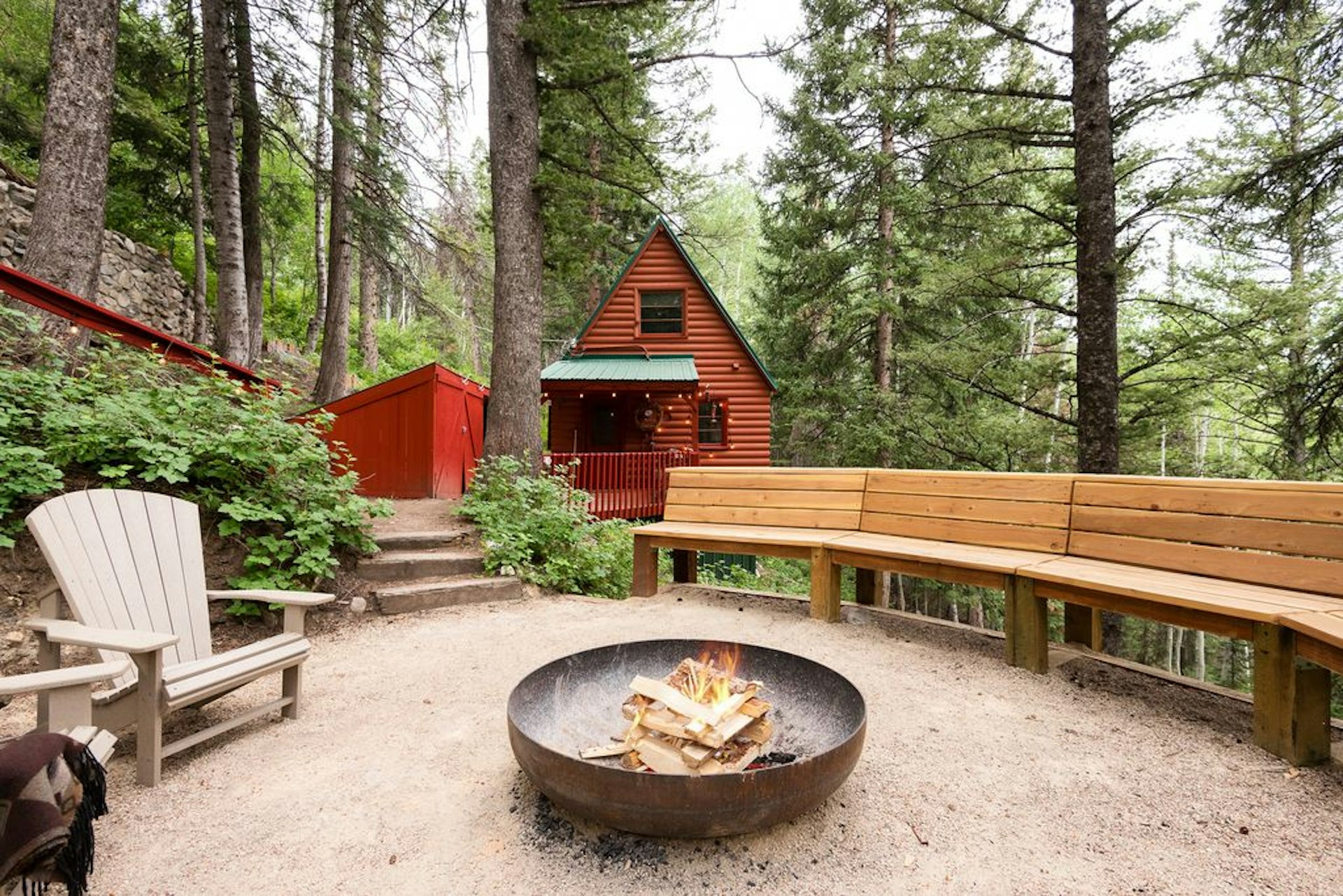 A red A-frame cabin surrounded by tall trees with a fire pit in the foreground