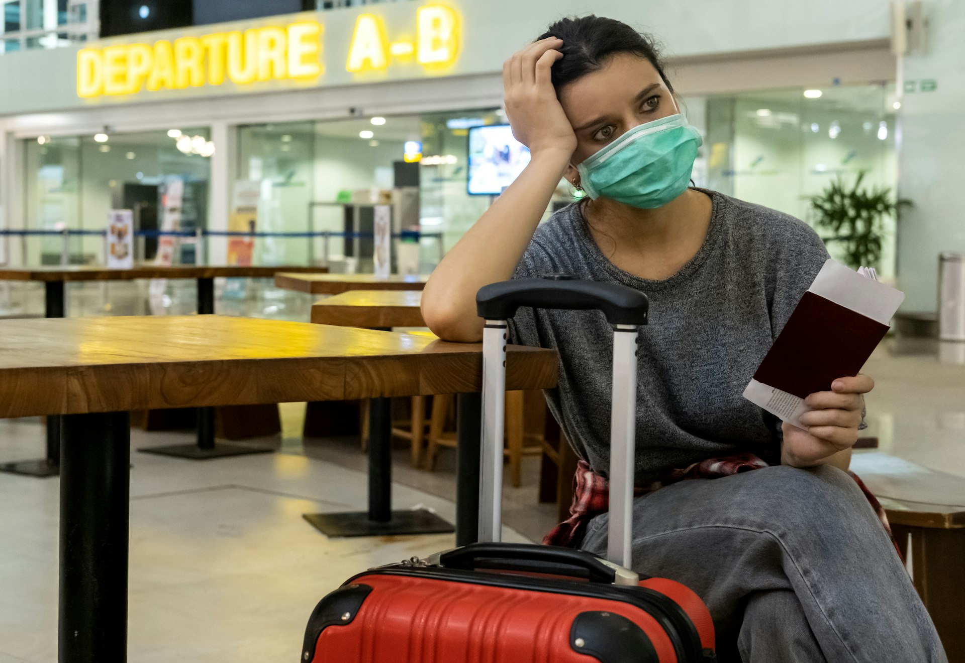 A female traveler wearing a green face mask sits alongside her luggage in the Departures area of an airport