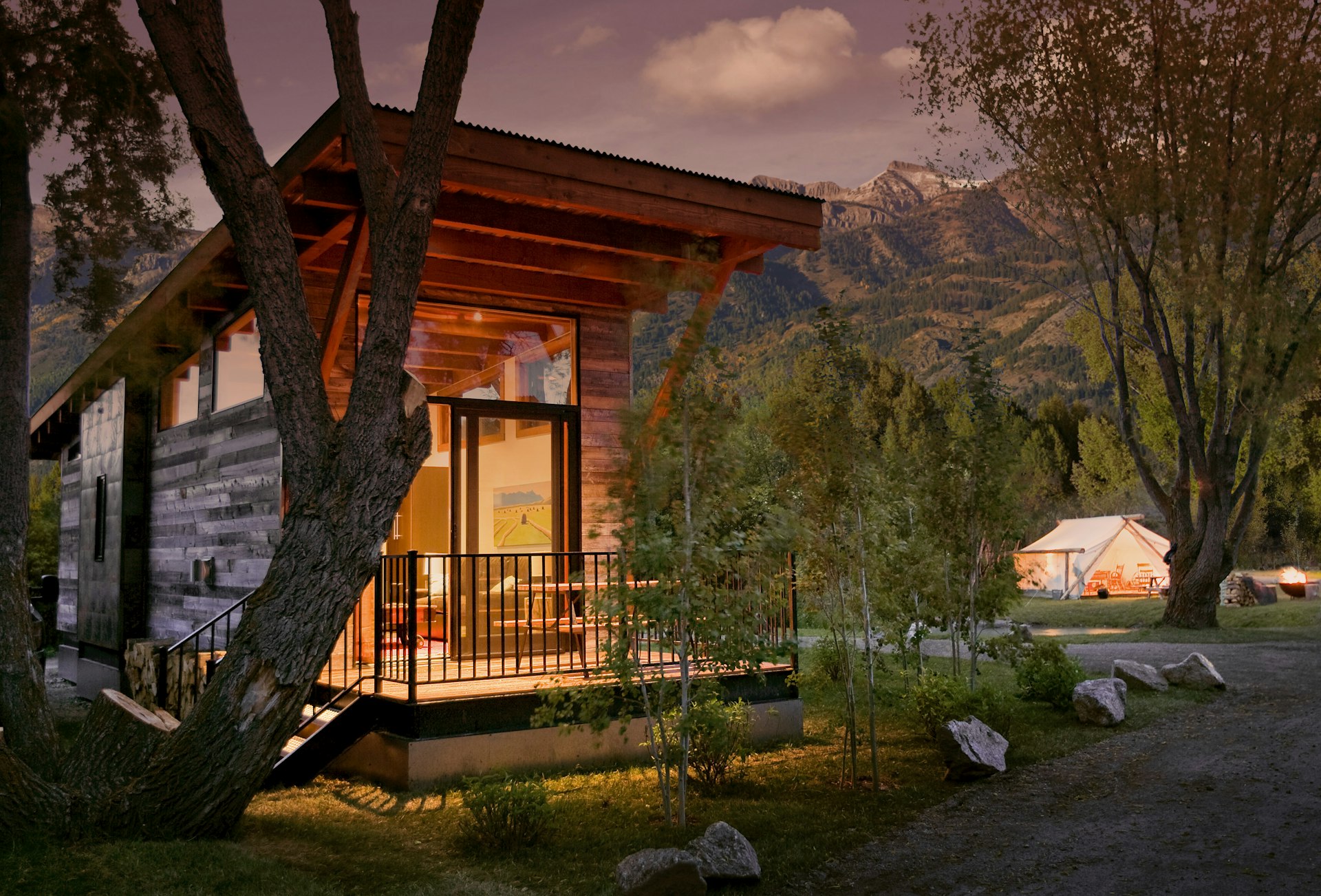 A stylish wooden cabin lit up at dusk. A large white tent is in the background. The mountains rise above the site in the distance.