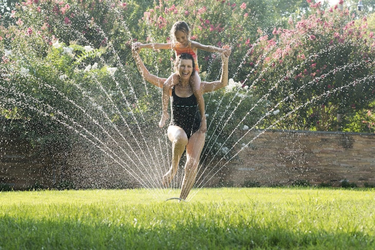 Mixed Race grandmother carrying granddaughter on shoulders in sprinkler