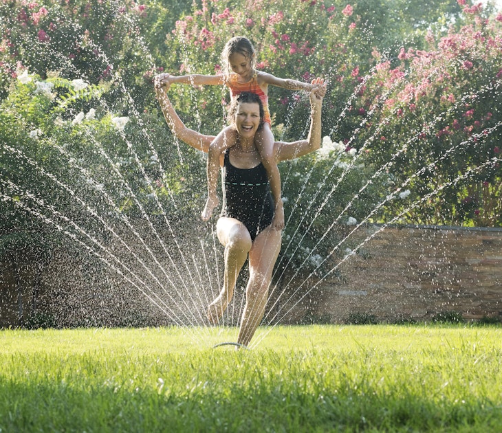 Mixed Race grandmother carrying granddaughter on shoulders in sprinkler