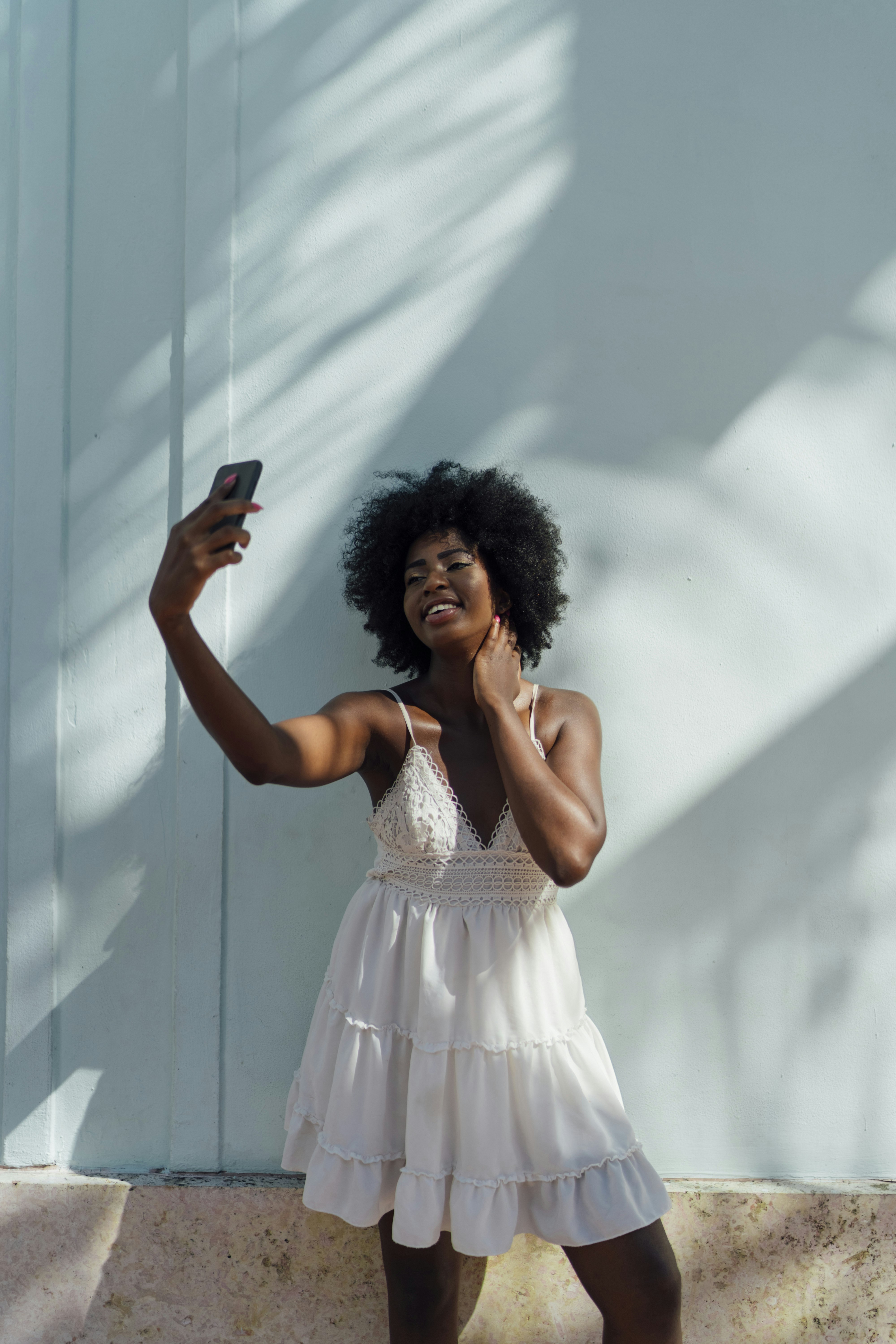 Smiling young woman wearing white dress taking a selfie at a wall