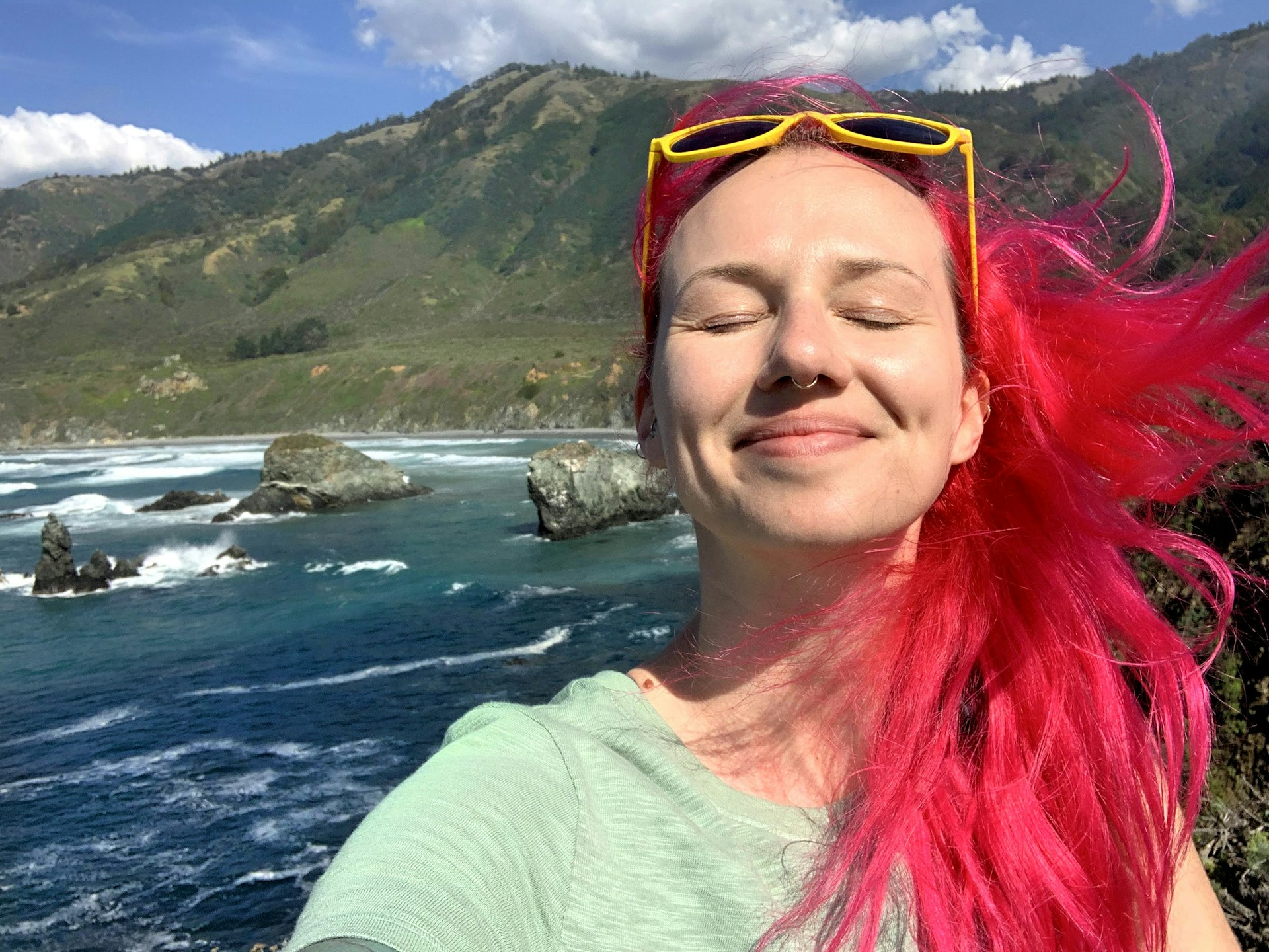 A woman with red hair smiling with her eyes closed with the Big Sur coastline behind her