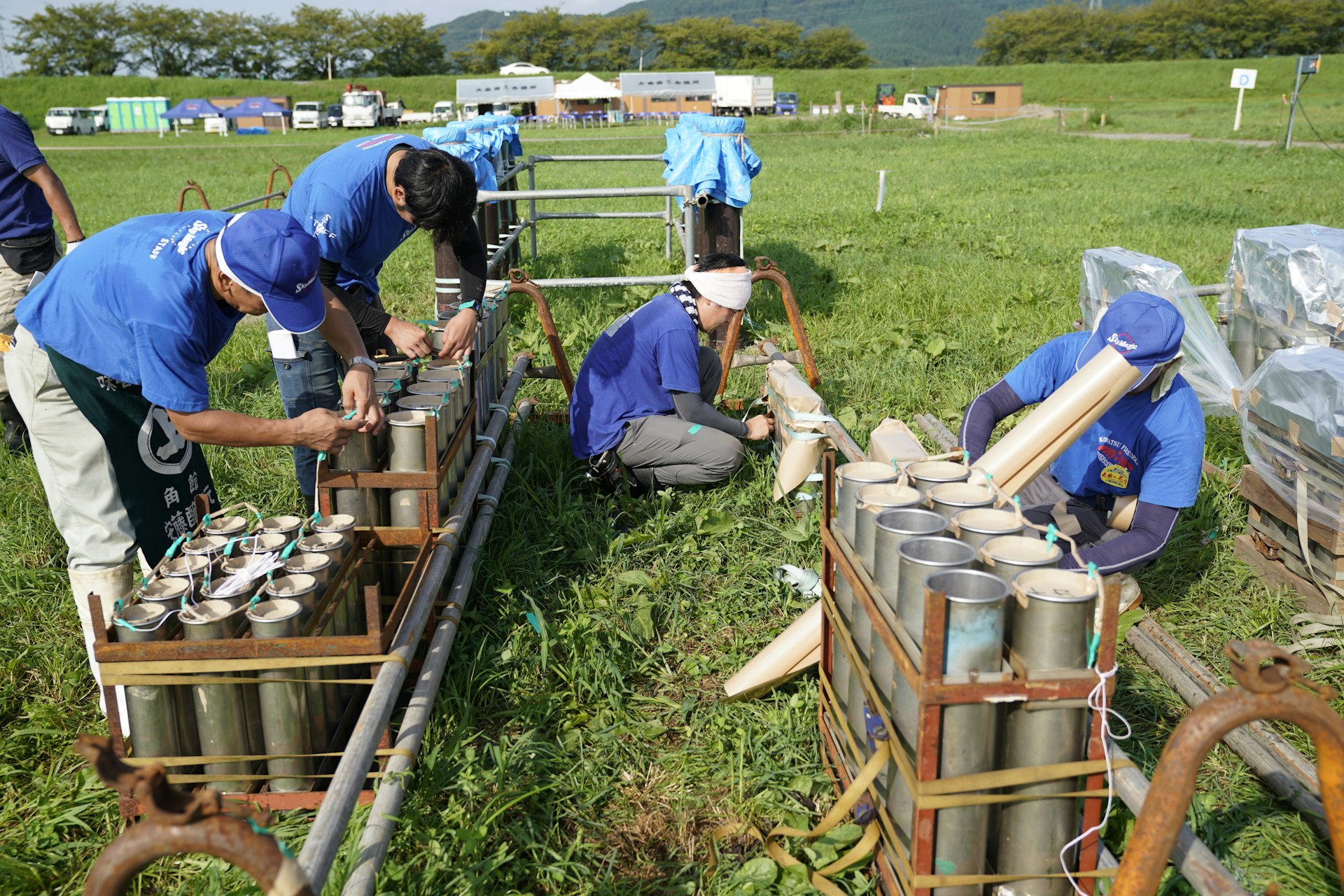 Four fireworks technicians bend over as they make adjustments to many tubes of explosives in an open field