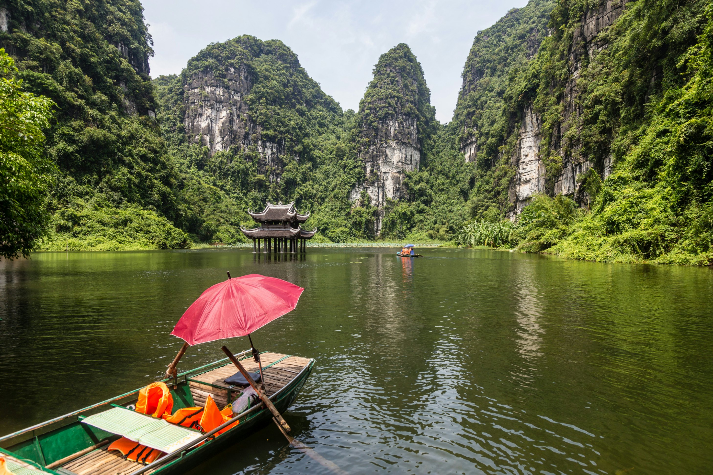Empty sampan with red umbrella waiting for tourists surrounded by steep limestone cliffs. Small temple in the water middle distance. Location of filming for King Kong