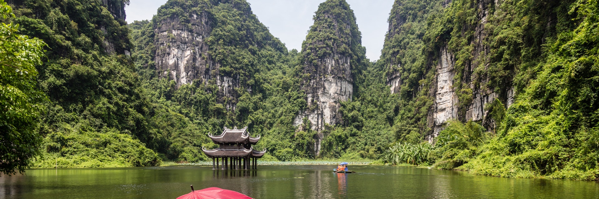 Empty sampan with red umbrella waiting for tourists surrounded by steep limestone cliffs. Small temple in the water middle distance. Location of filming for King Kong