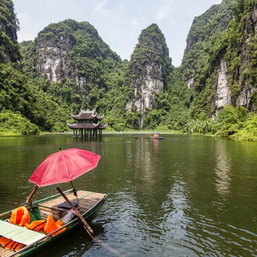 Empty sampan with red umbrella waiting for tourists surrounded by steep limestone cliffs. Small temple in the water middle distance. Location of filming for King Kong