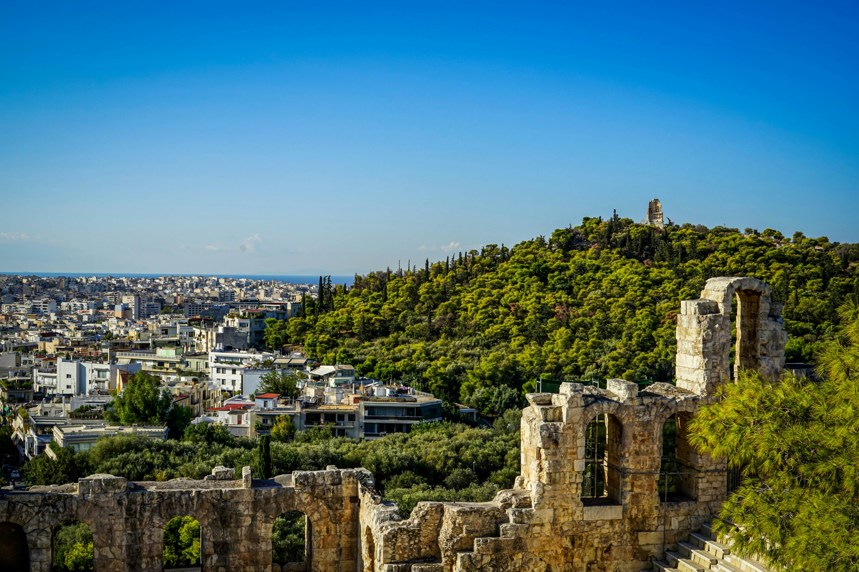 View of Athens cityscape and the Philopappu Monument through ancient stone theatre seeing lowrise white buildings architecture, mountain, trees and clear blue sky background, Greece
