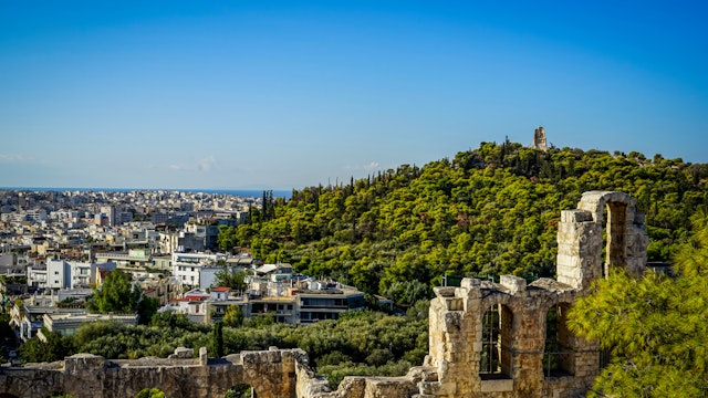 View of Athens cityscape and the Philopappu Monument through ancient stone theatre seeing lowrise white buildings architecture, mountain, trees and clear blue sky background, Greece