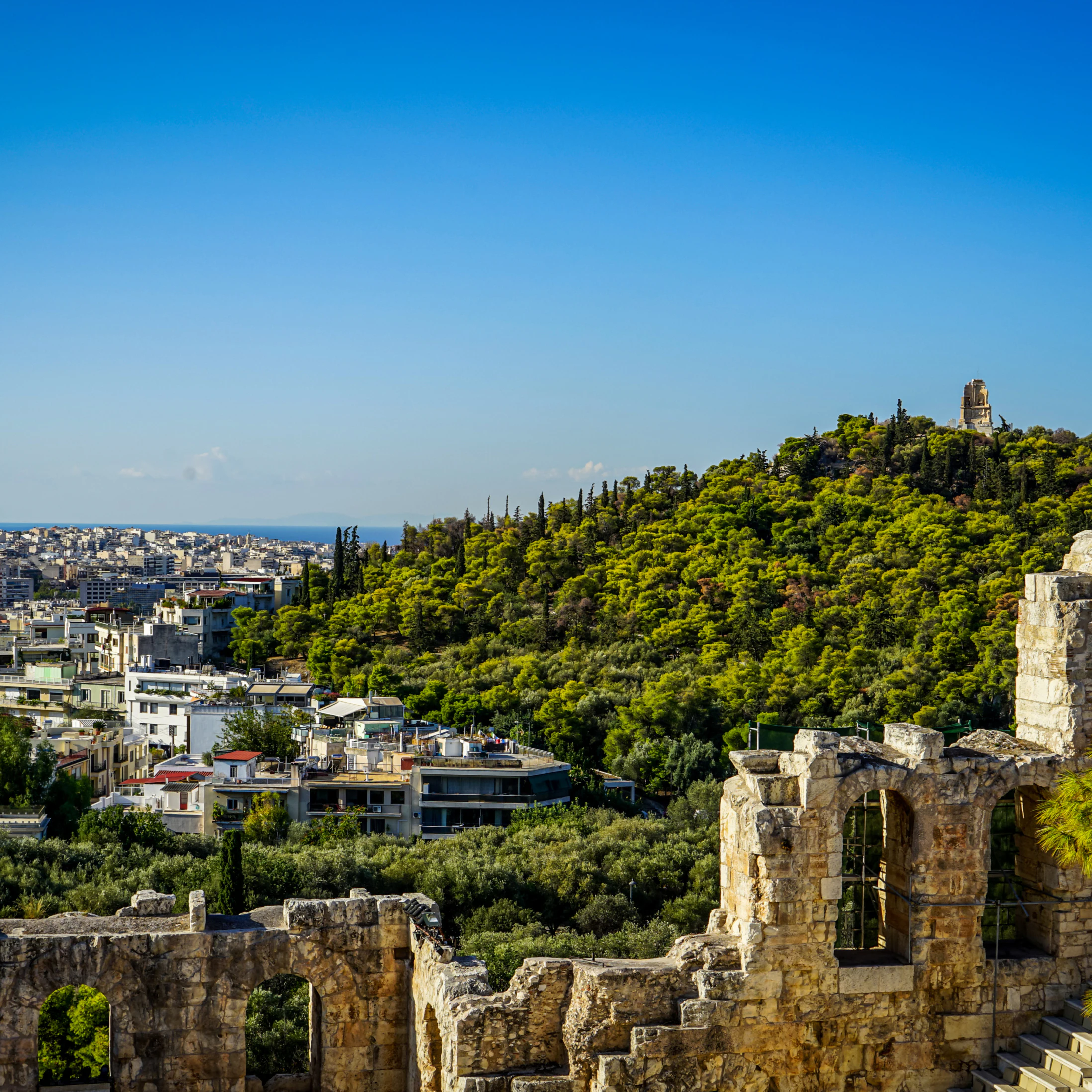 View of Athens cityscape and the Philopappu Monument through ancient stone theatre seeing lowrise white buildings architecture, mountain, trees and clear blue sky background, Greece