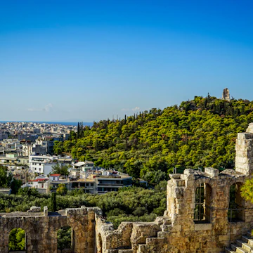 View of Athens cityscape and the Philopappu Monument through ancient stone theatre seeing lowrise white buildings architecture, mountain, trees and clear blue sky background, Greece