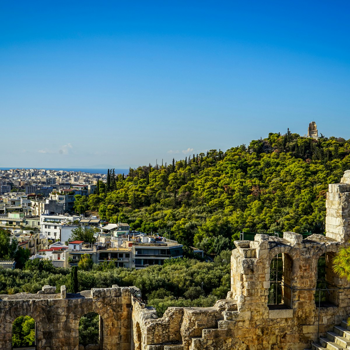 View of Athens cityscape and the Philopappu Monument through ancient stone theatre seeing lowrise white buildings architecture, mountain, trees and clear blue sky background, Greece