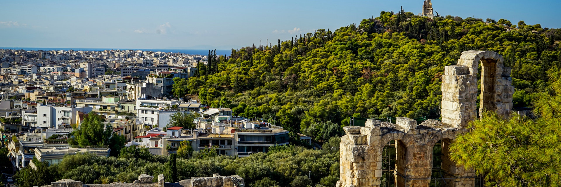 View of Athens cityscape and the Philopappu Monument through ancient stone theatre seeing lowrise white buildings architecture, mountain, trees and clear blue sky background, Greece
