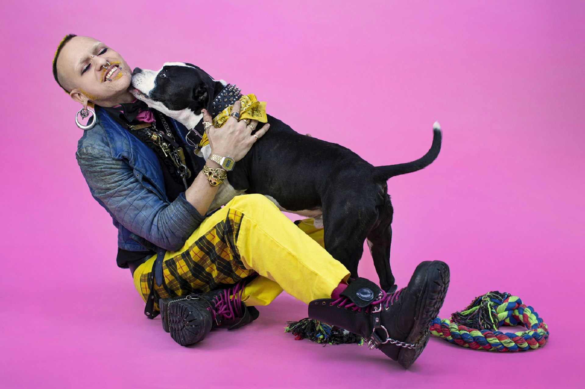 A non-binary person dressed in punk style is being licked on the face by their black-and-white dog