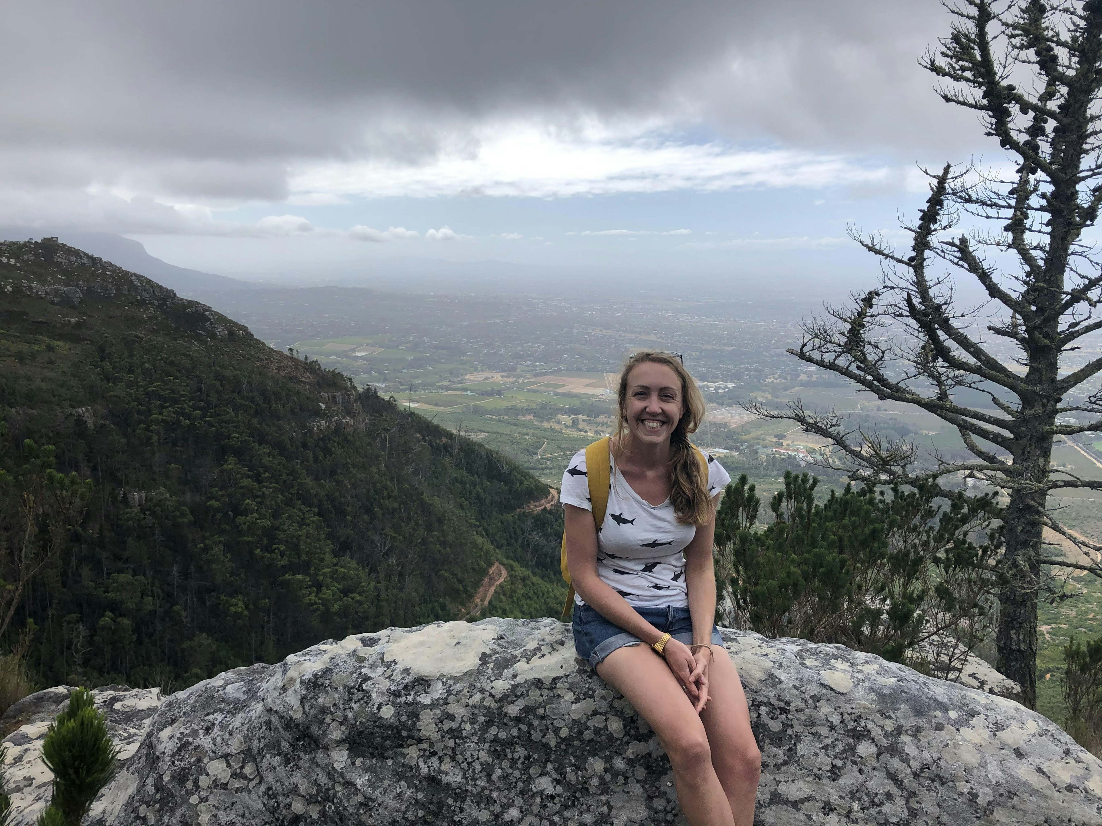 A woman sits on a rock in dense countryside, smiling cheerily at the camera