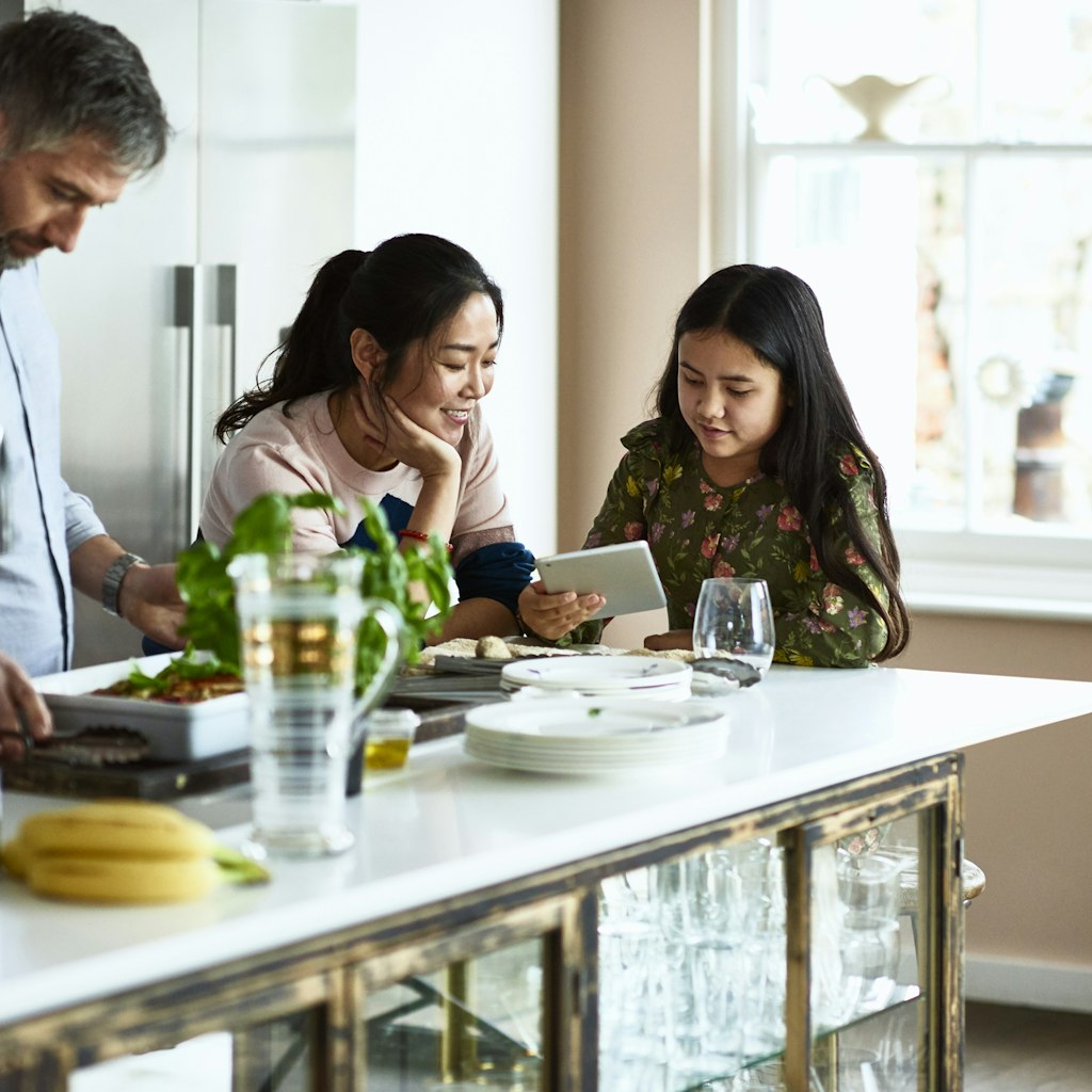 Modern family life with mature man making dinner, wife and mixed race girl using device, sitting around kitchen island, freshly home cooked meal on work surface