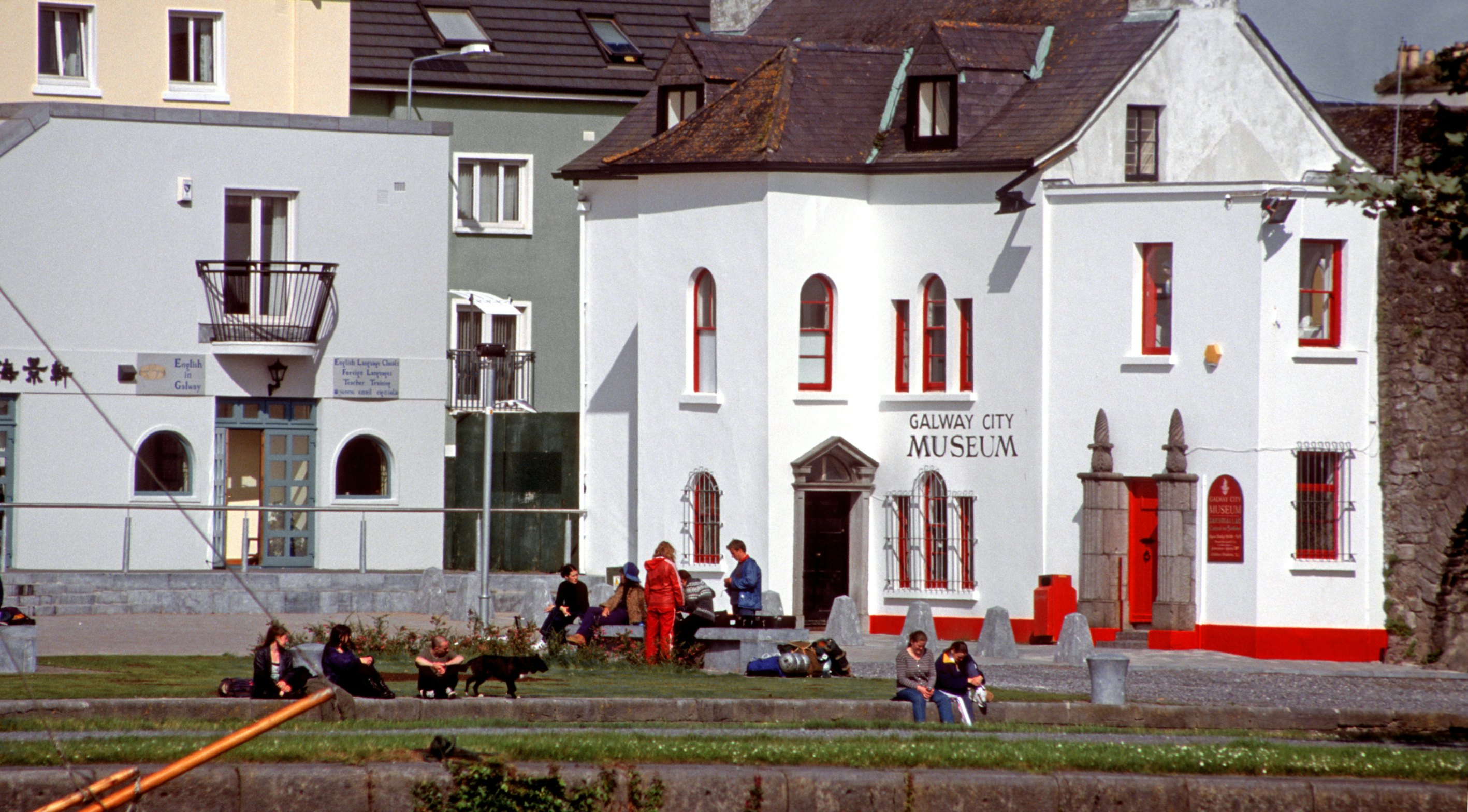 Galway, Ireland - June 17th 2005: Teenagers, some with backpacks, sitting on the bank of river Corrib, front the Galway City Museum.