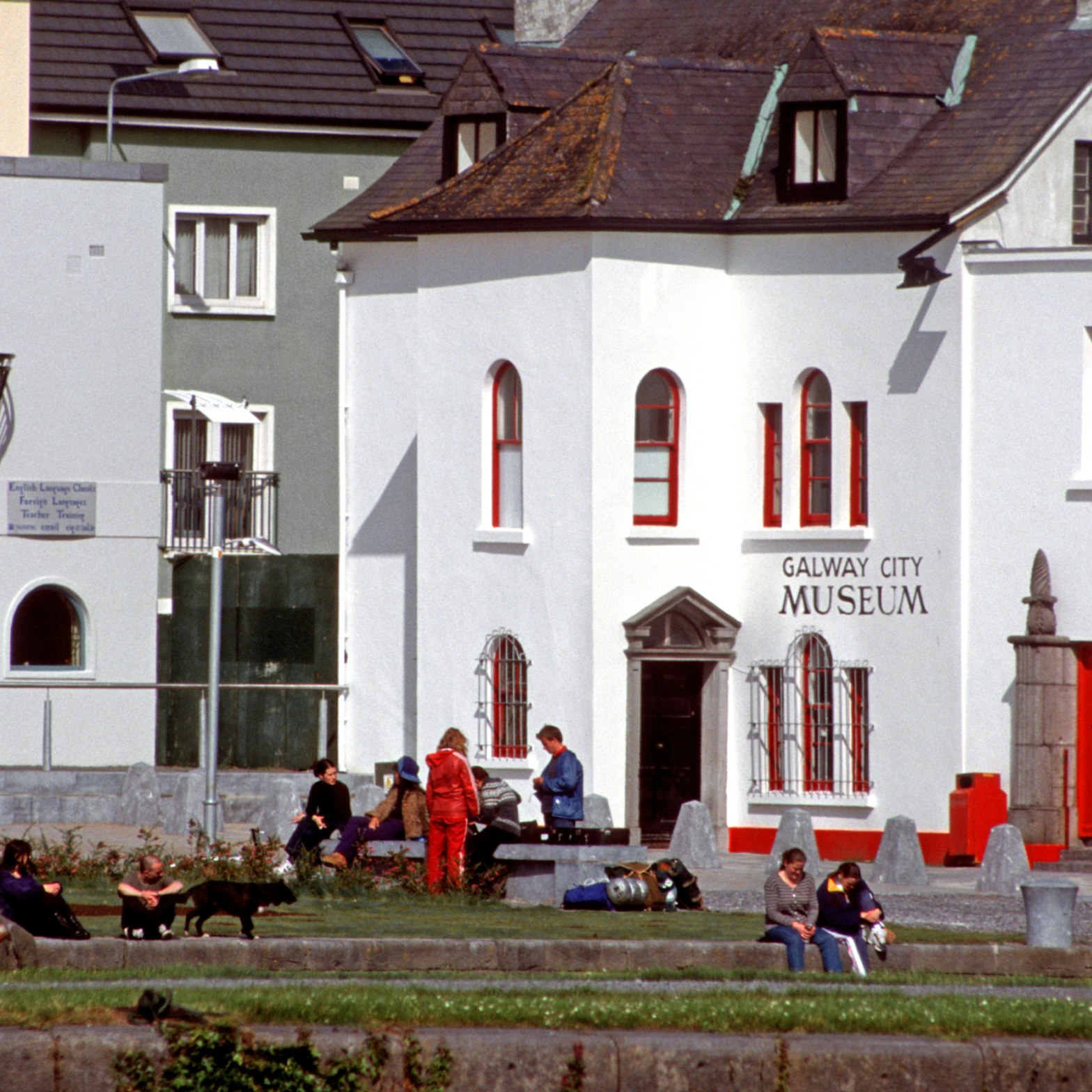 Galway, Ireland - June 17th 2005: Teenagers, some with backpacks, sitting on the bank of river Corrib, front the Galway City Museum.