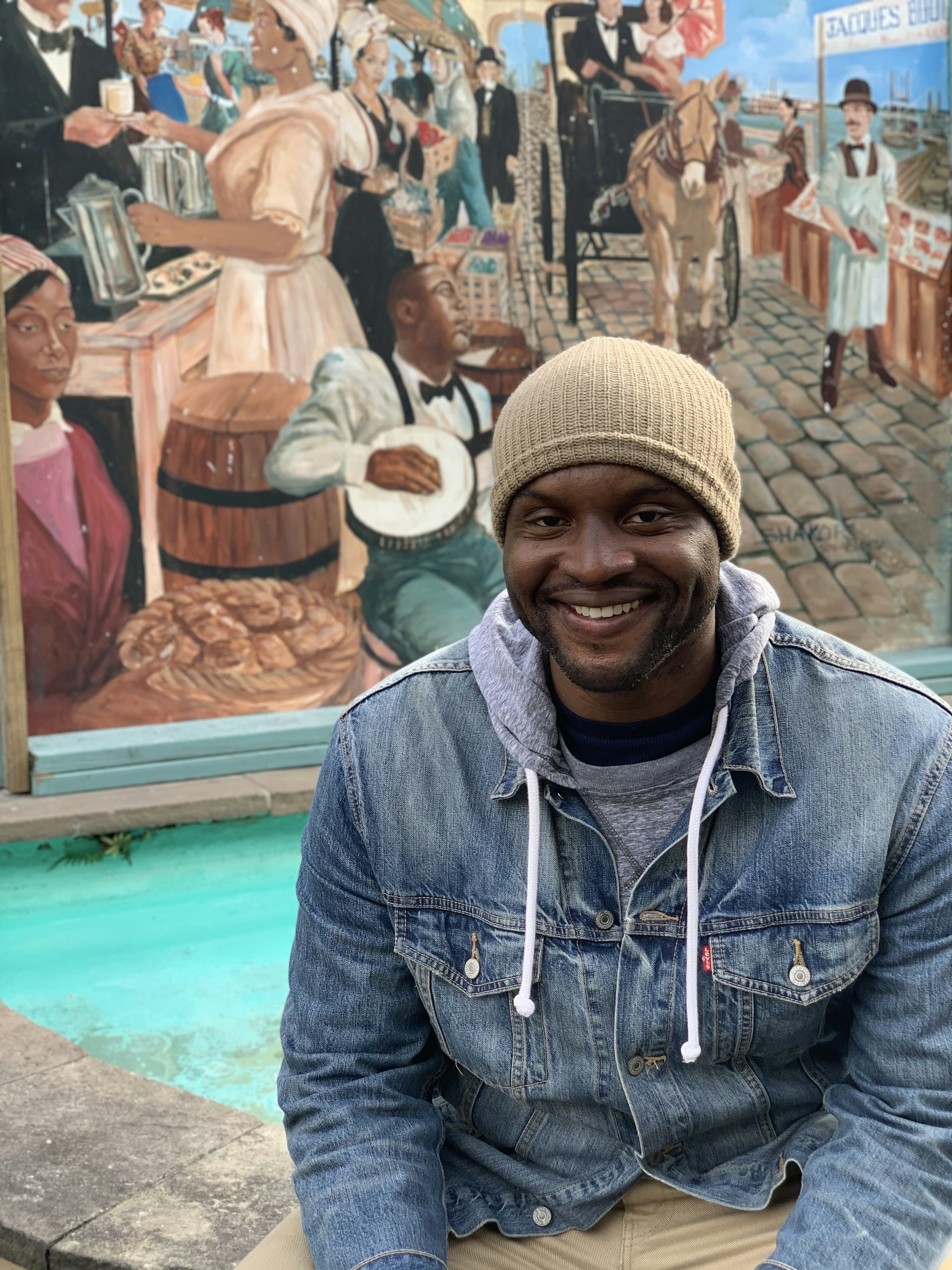 A black man sitting in front of a mural smiles at the camera