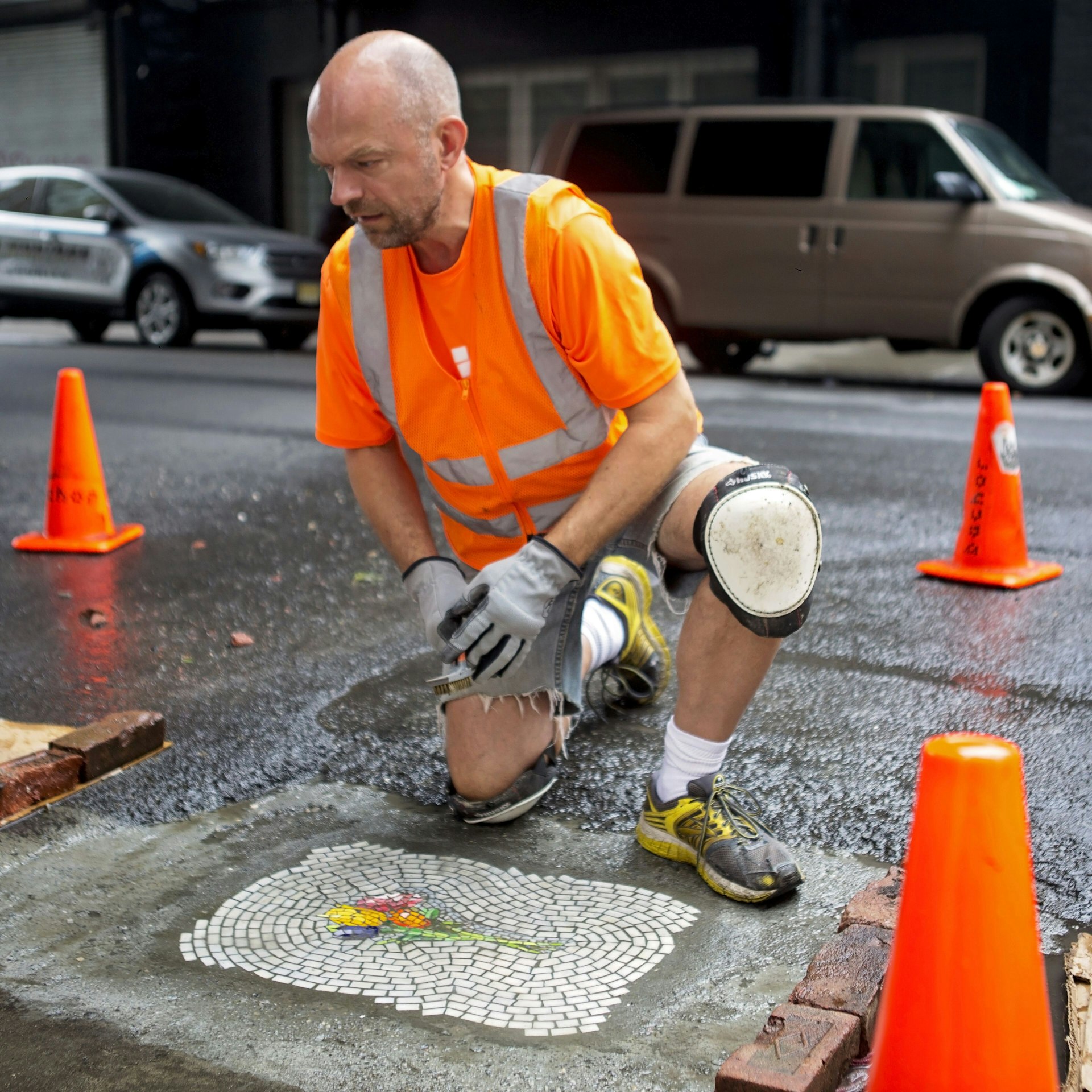 Jim Bachor wears an safety orange t-shirt and reflective vest as he crouches between three orange traffic cones in gloves and kneepads over a white mosaic with a cluster of colorful flowers at its center