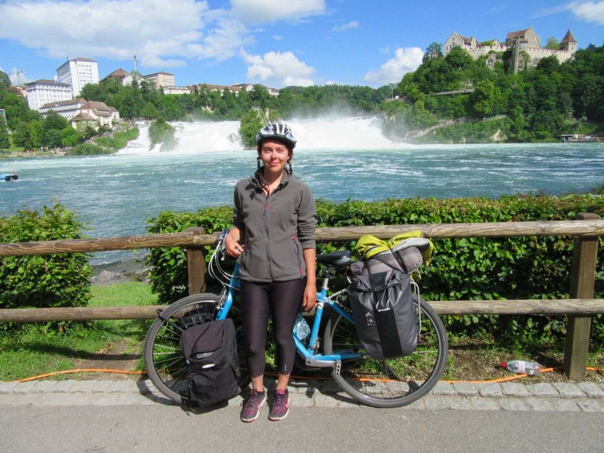 A woman stands with her bike in front of a cascading waterfall with a castle on the hill to the right