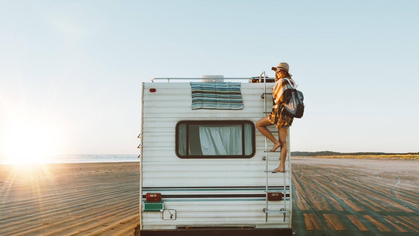 Young woman on the back of an RV, which is parked on the beach during sunset.