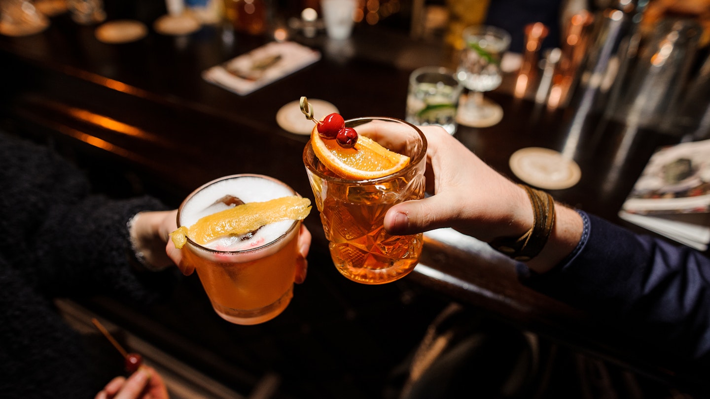 A man and a woman cheersing glasses at a bar counter - a sour mix and Negroni with orange and cherry.