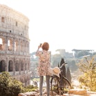 Beautiful young woman in colorful fashion dress alone on a hill with bike takes pictures of colosseum in Rome using smartphone camera at sunset. Attractive tourist girl with elegant straw hat.