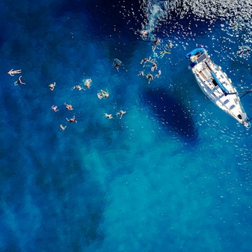 Aerial of a group of people swimming in blue water near a sailing boat in Barbados.