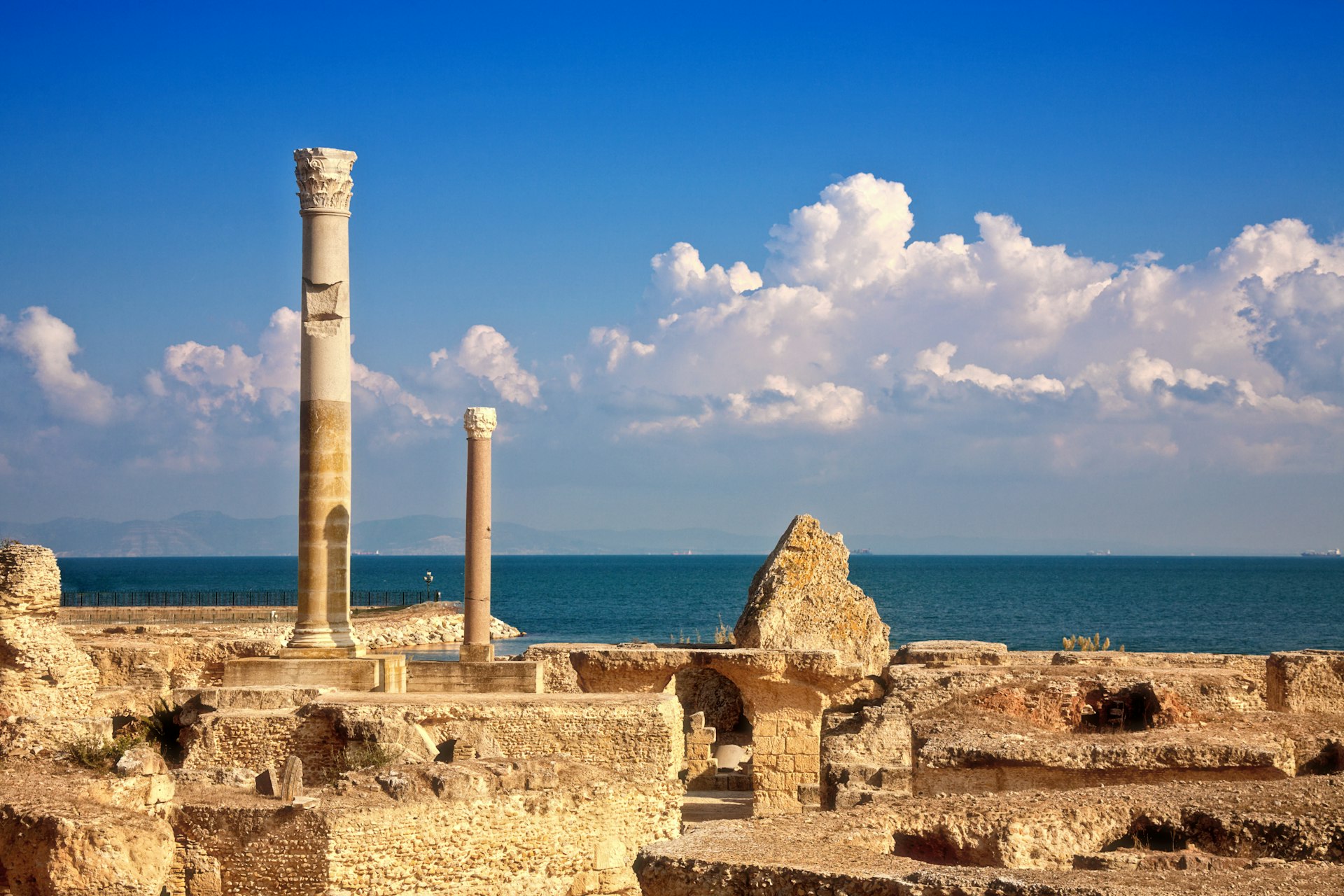 Ruins, dominated by a free-standing chimney-like structure. The blue sea stretches out in the distance