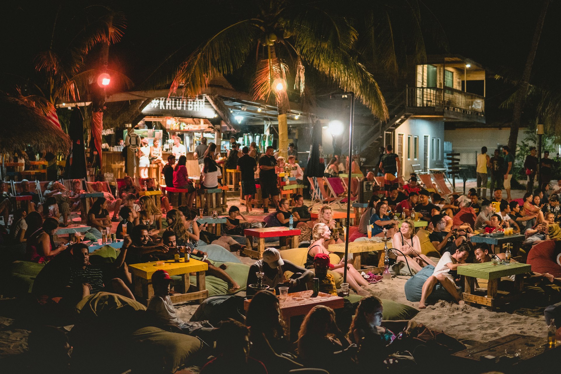 Large numbers of revelers sit on the sand and at tables drinking cocktails under overhead lights on Cenang Beach