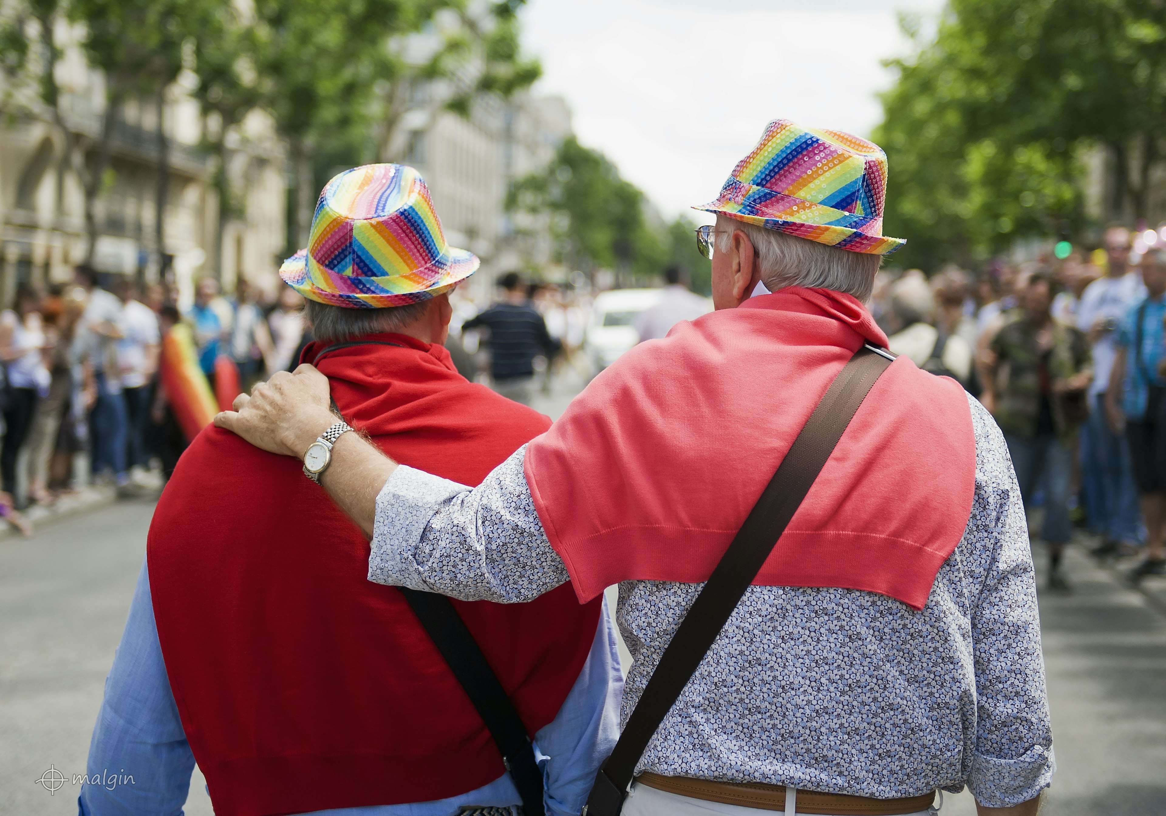 Two men viewed from behind. They're walking along together, one with his arm around the other. Both have red jumpers slung over their shoulders and are wearing rainbow hats