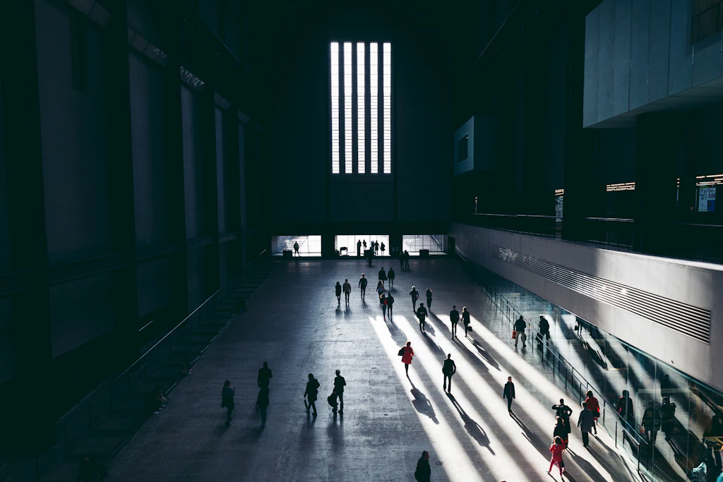 A shadowy huge space lit by large but narrow windows. People with long shadows are walking along the concrete floor