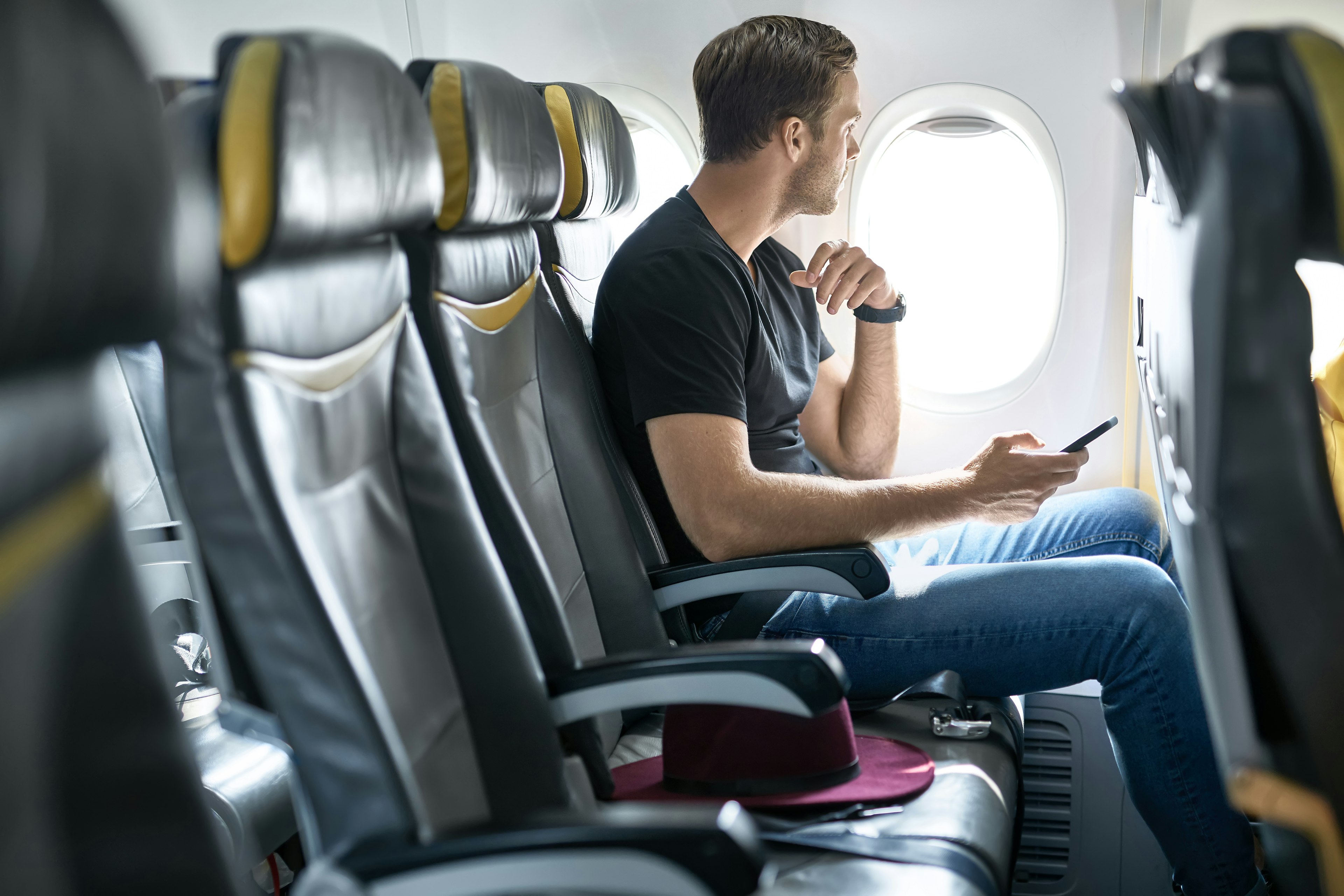 A young male passenger sits in an airplane seat next to the window with a mobile phone