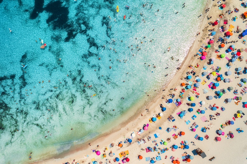 Aerial of a crowded sandy beach with colourful umbrellas, sun bathers and swimmers during summer.