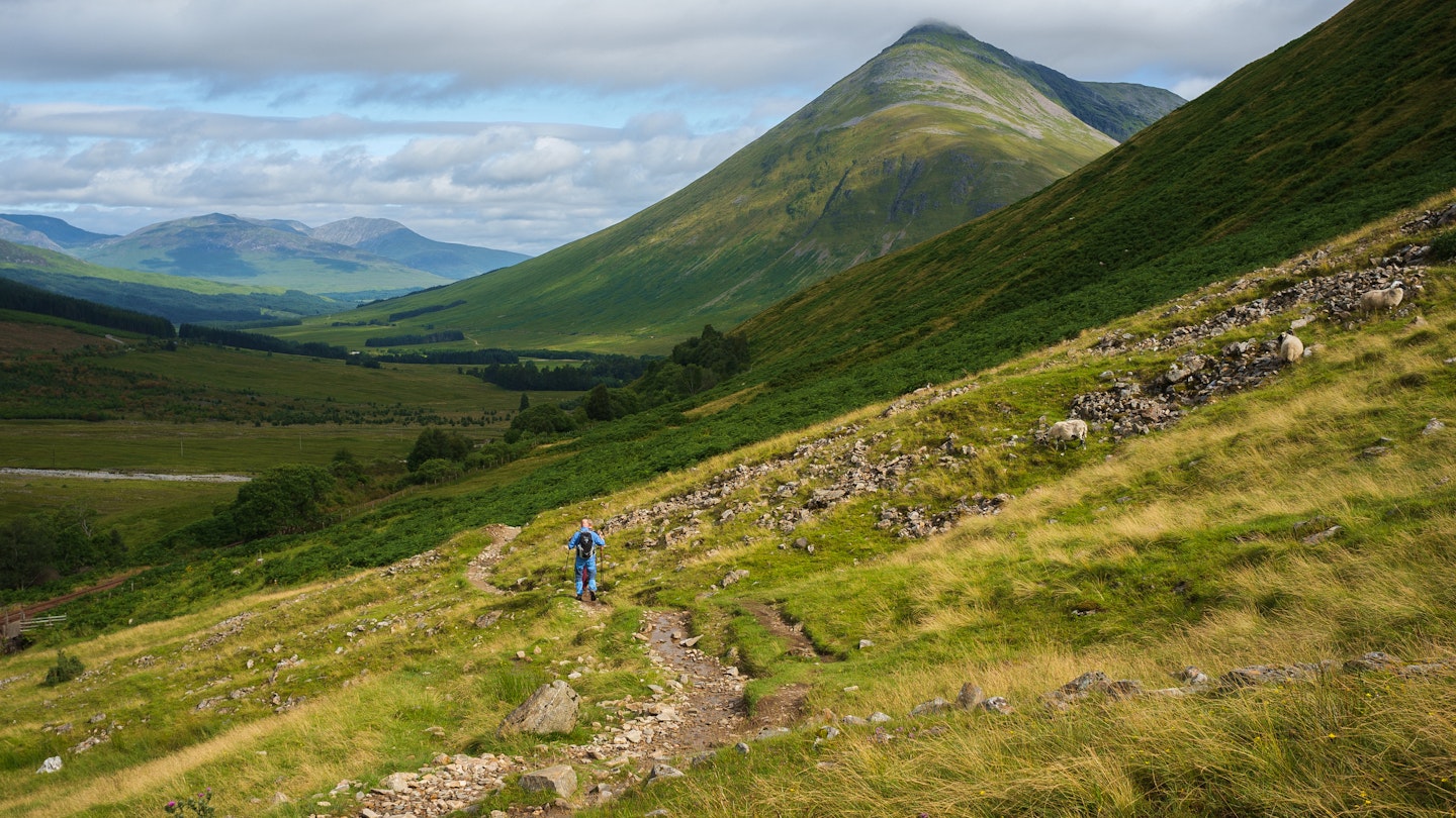 Two people hiking on a rocky trail through grassy hills in the Scottish highlands.