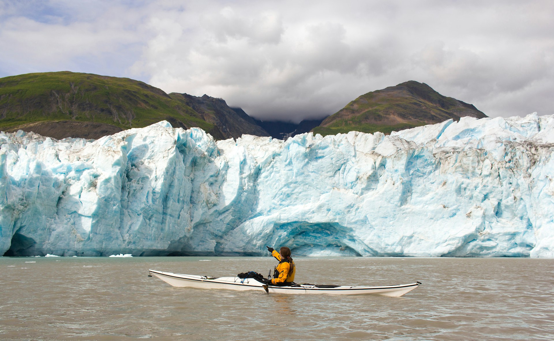 A person wearing a yellow jacket in a white kayak points at a vast wall of ice with green hills behind