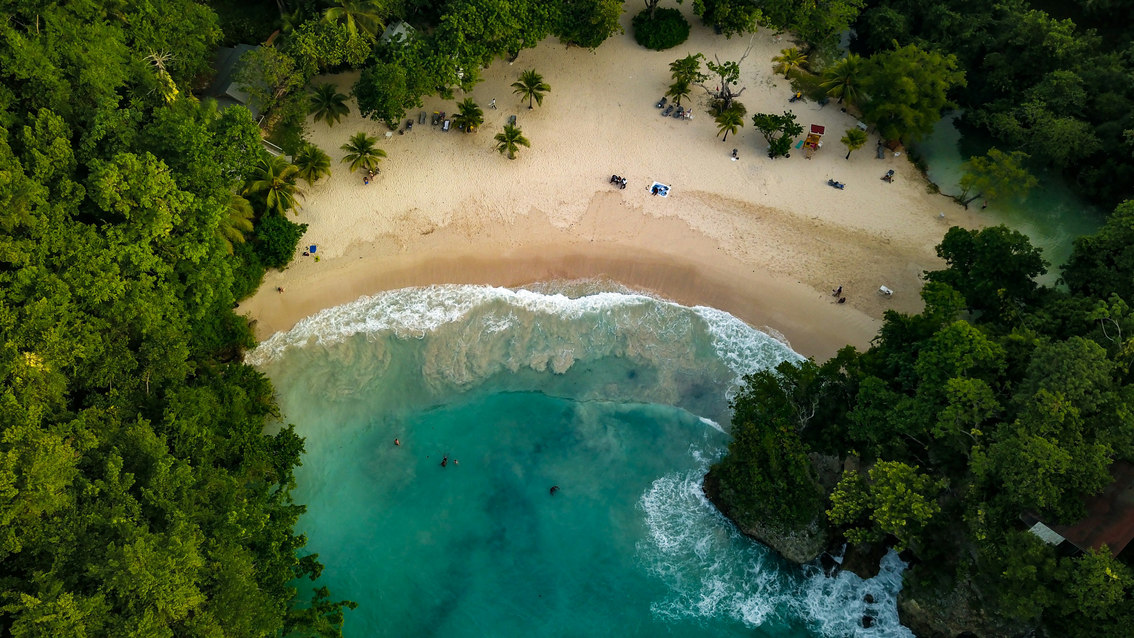 Aerial of waves crashing on the shore at Frenchman's Cove in Jamaica. There are groups of people scattered around the beach.