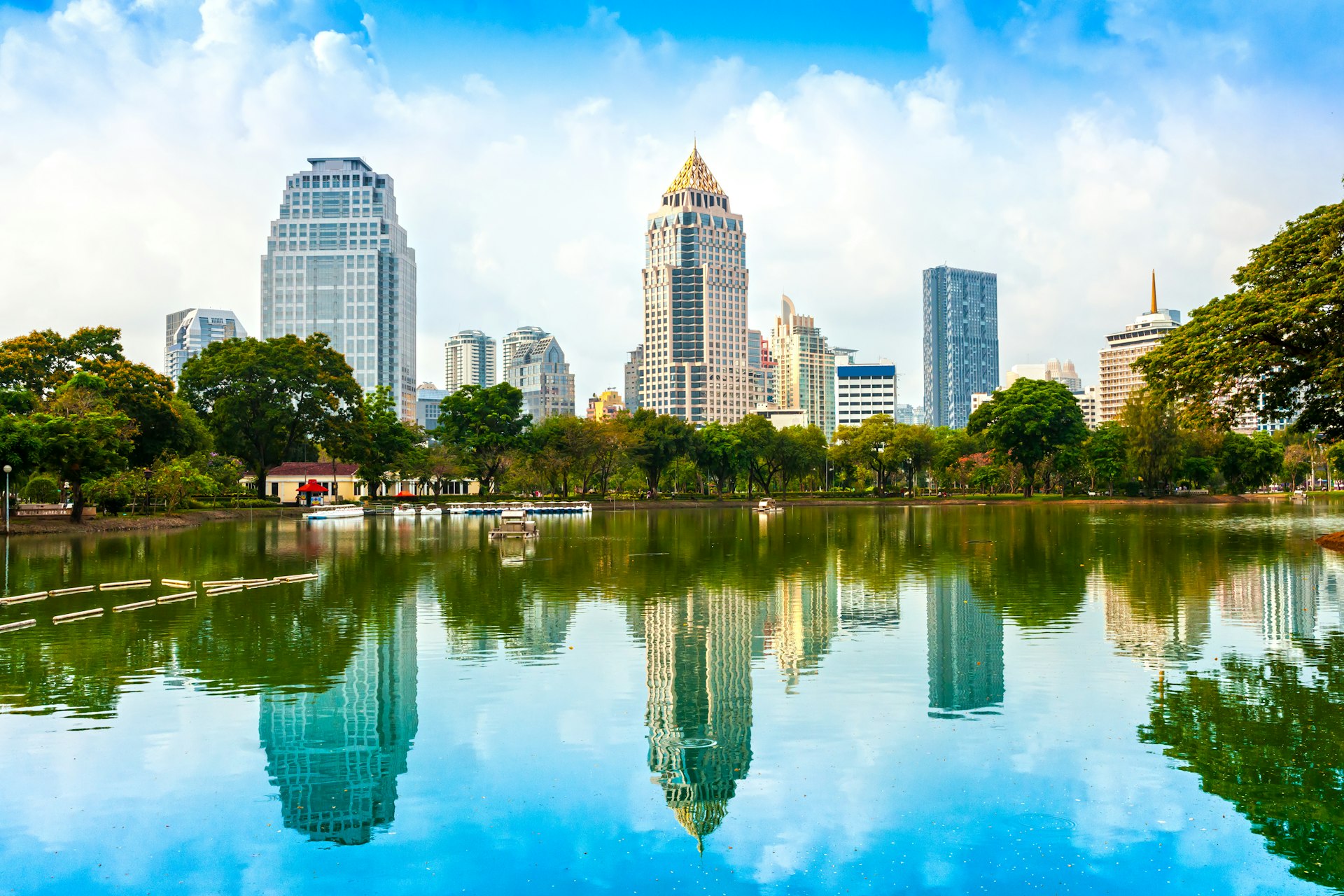 The tall skyscrapers of the Bangkok skyline sit in front of a bright blue sky across the glittering, quiet and empty lake that belongs to Lumphini Park