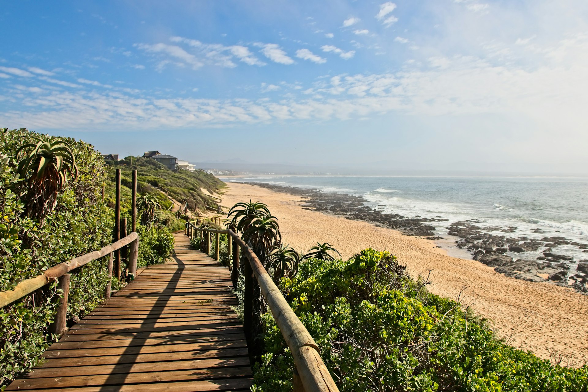 A plant-lined boardwalk leading down to a golden sand beach with some rocks near the sea